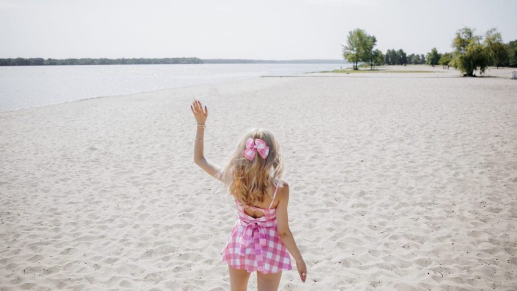 Young blonde girl look like a fashion doll doll in pink checkered mini dress and pink bow in her hair waving her hand on beach against sand and water background. Back view. Close-up.