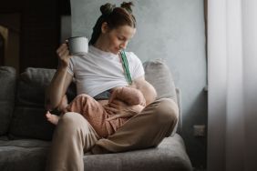 Young mother breastfeeding her baby while drinking beverage from a cup.