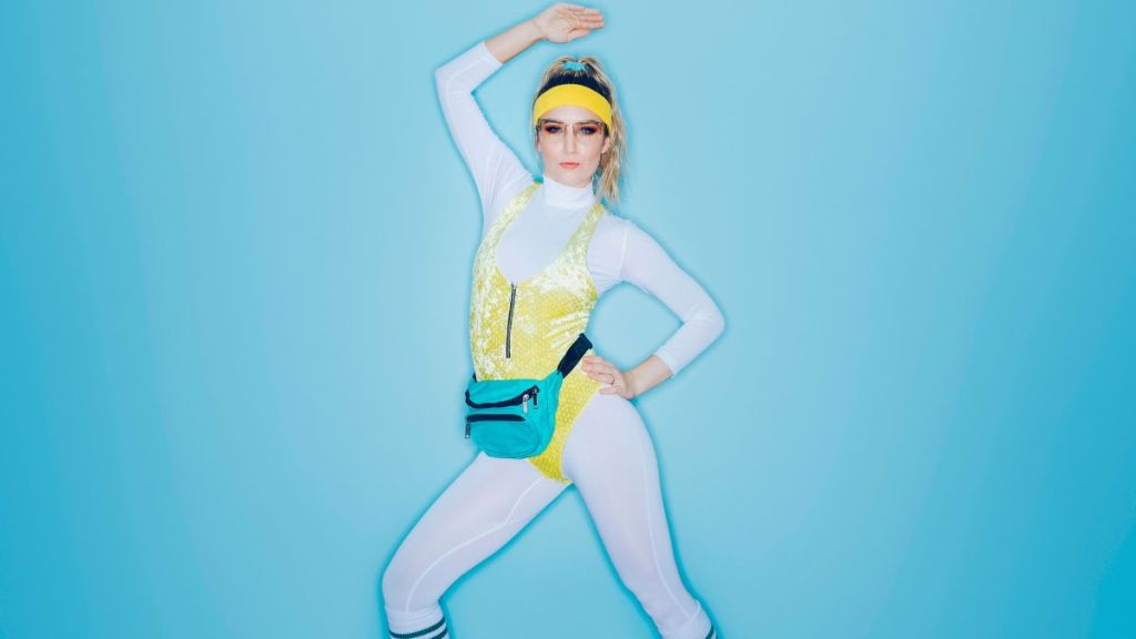 A woman wearing exercise clothing styled after the 1980's and 1990's does aerobics style exercises in front of a large bright blue background.