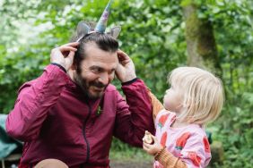 Little girl looks up at her dad as he tries on her unicorn headband and tries to make her laugh.