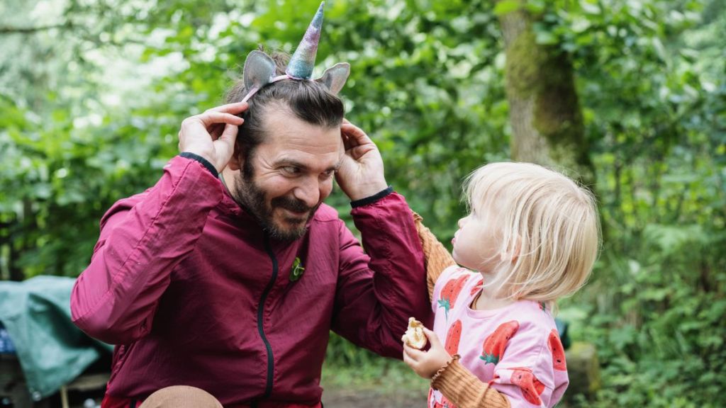 Little girl looks up at her dad as he tries on her unicorn headband and tries to make her laugh.