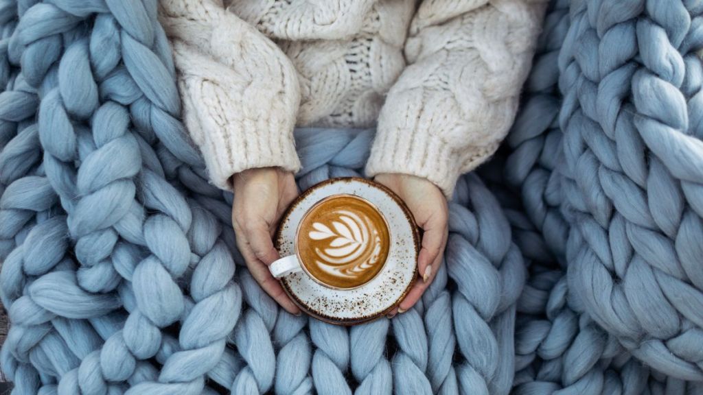 Female hands in white woolen sweater holding cup of hot butterscotch latte covered with woolen merino blanket.