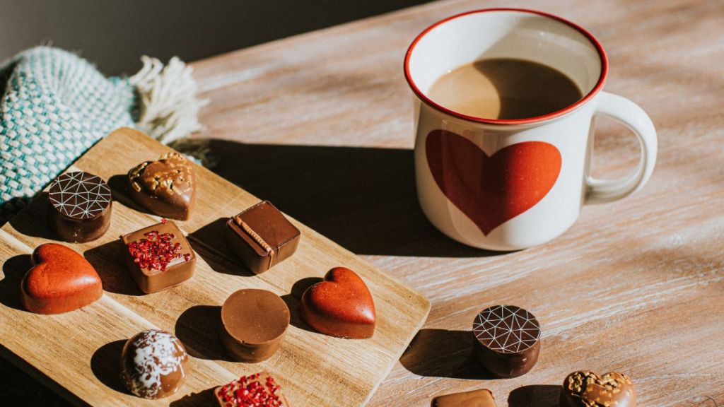 Luxury chocolates in various shapes and flavours, including red heart chocolates beside a hot mug of coffee with a large heart emblem, perfect for Valentine's Day gifting.