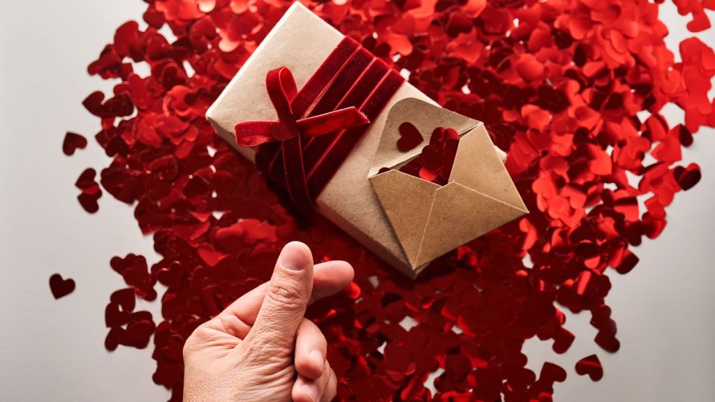 Craft gift and envelope with many red shiny hearts. Hand with thumb and index finger shows heart symbol in asia. A wonderful photo for Valentine's Day.