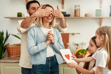 A young girl surprises her mom with a handmade Valentine's Day card while her father holds her eyes closed.