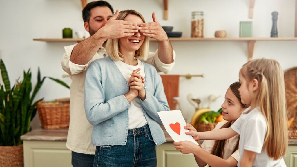 A young girl surprises her mom with a handmade Valentine's Day card while her father holds her eyes closed.