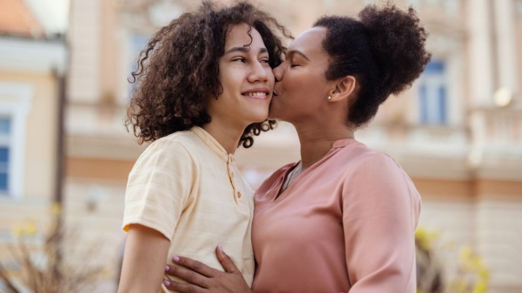 A Tender Moment of an Affectionate Mother Kissing Her Teenage Son on the Cheek Outdoors, Showcasing Their Strong Bond and Mutual Love.