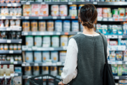 Rear view of young Asian mother with a shopping cart grocery shopping for baby products in a supermarket. She is standing in front of the baby product aisle and have no idea which product to choose from